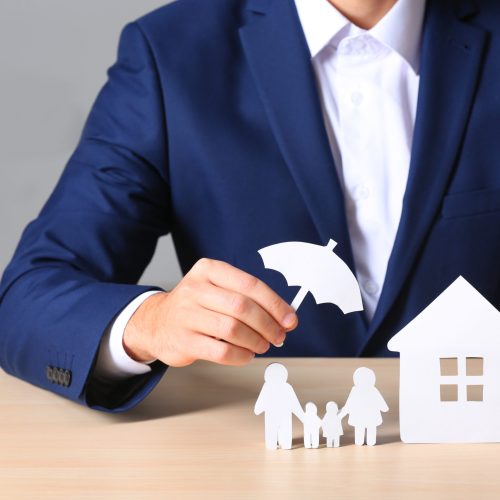 Male insurance agent covering paper family and home with umbrella cutout at table, closeup
